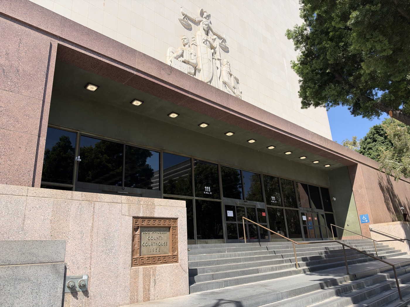 The Hill Street entrance of the Stanley Mosk Courthouse at 111 North Hill Street, with the figure of Justice holding scales above the doors and a cornerstone reading Los Angeles County Courthouse 1958